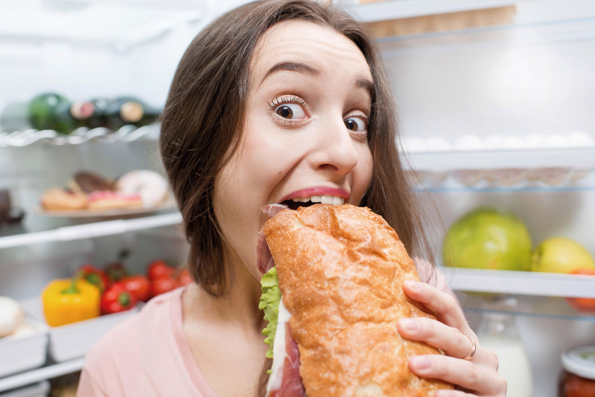 Young woman eating big sandwich in front of the refrigerator full of friuts and vegetables