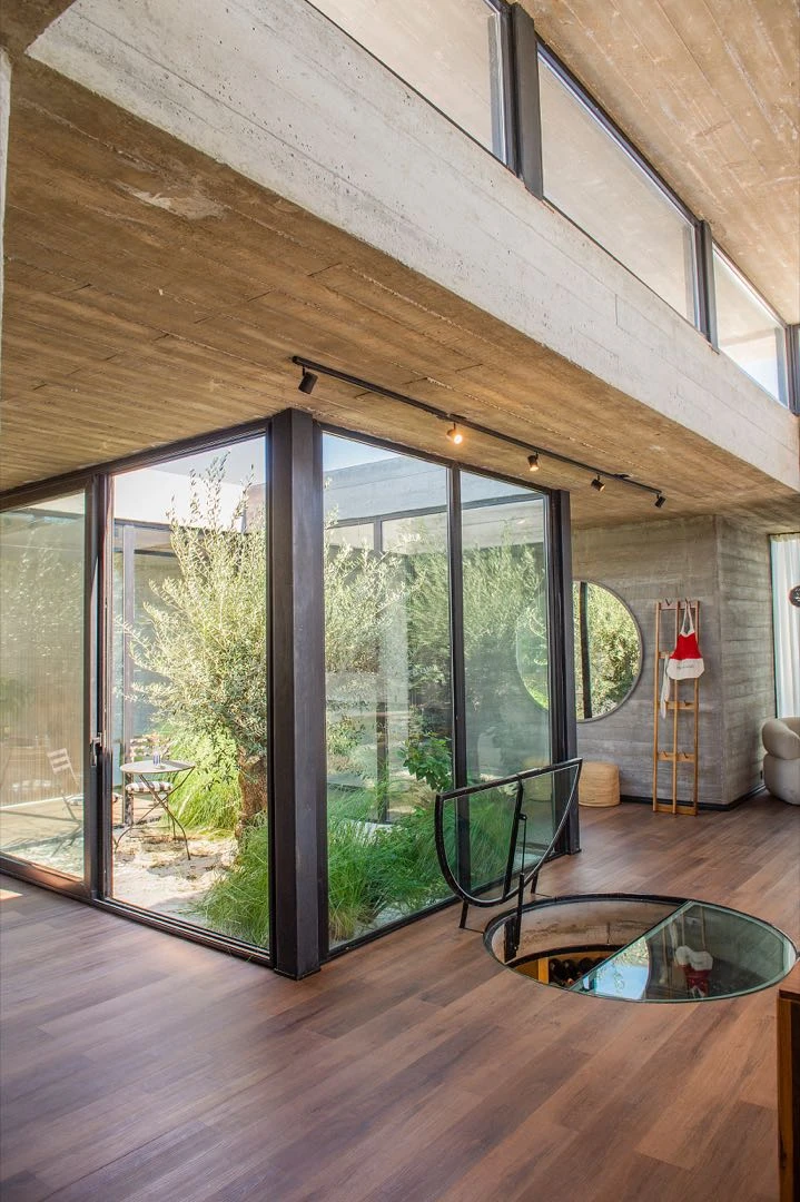 El acceso a la cava, calado en el piso de madera del living comedor. El patio interno baña al espacio de luz y verde.