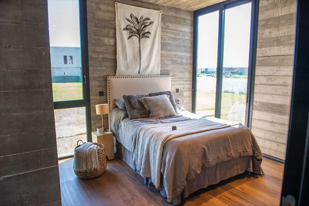 Un dormitorio alternativo con paredes de hormigón y ventana corrediza de piso a techo con vista al lago. 