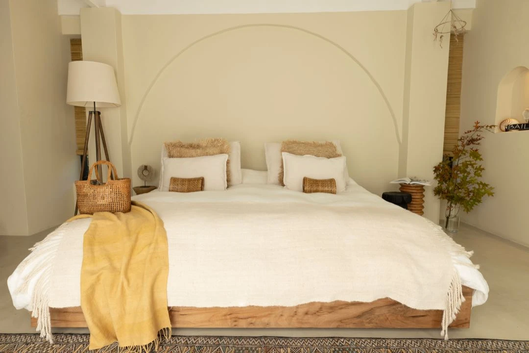 La cama king size con base de madera al natural se viste con ropa de cama de lino en tonos crudos con detalles en ocre y fibras naturales. 