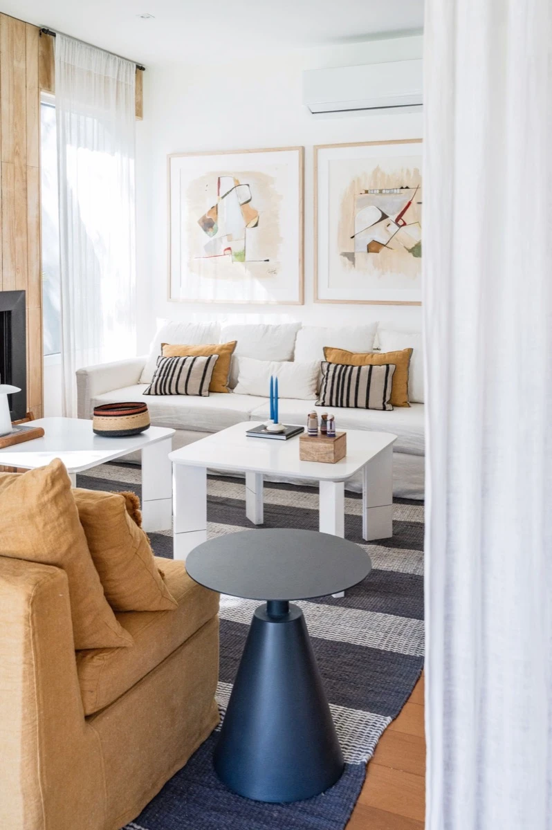 Sofá blanco y almohadones en color mango y rayados (Delfina Constantini). Los cuadros continúan el lenguaje artístico. 