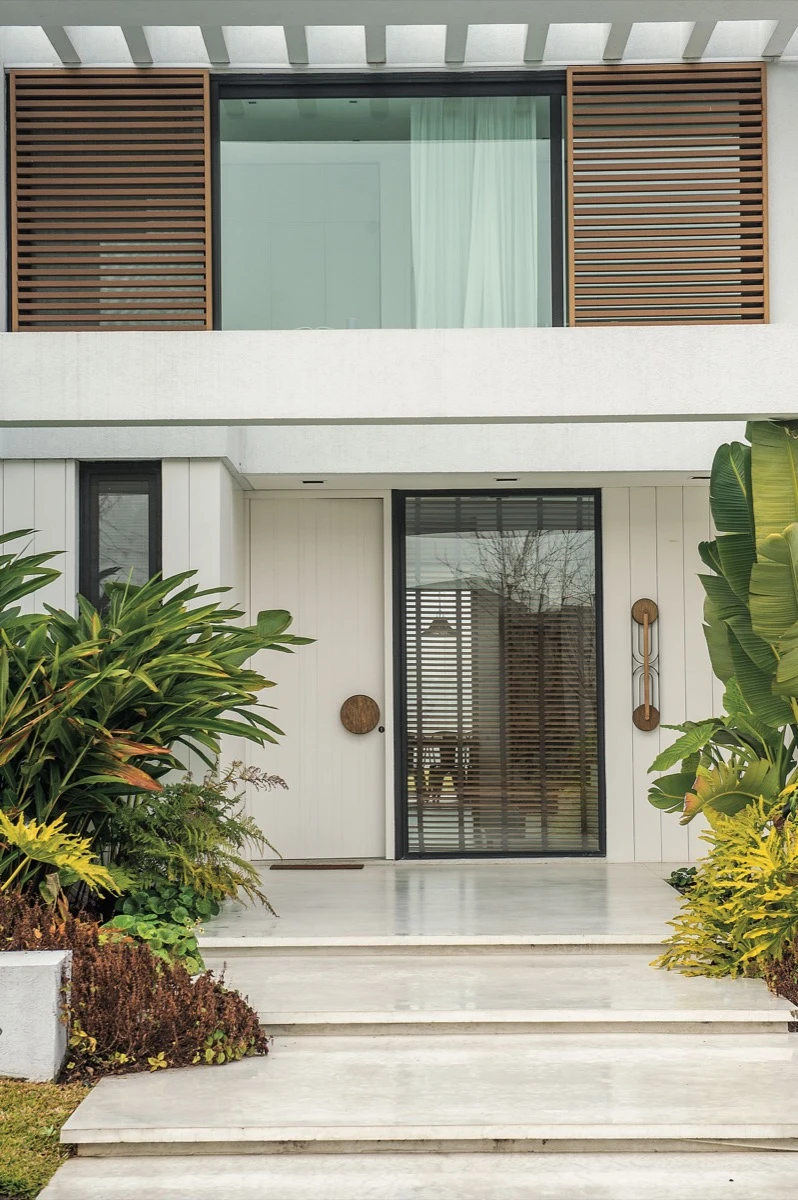 El ingreso a la casa se da a través de unos escalones de cemento alisado, con dos canteros laterales llenos de plantas tropicales,