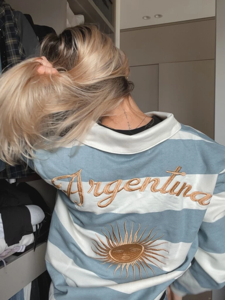 El diseño se distingue por su cuello tipo polo blanco, su silueta amplia y un detalle que marca la diferencia: en la espalda lleva la inscripción “Argentina” bordada en hilo dorado con tipografía cursiva, acompañada por un Sol de Mayo también bordado. 