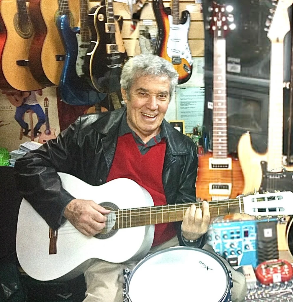 En la imagen, Chico Novarro aparece sonriente dentro de un local de instrumentos musicales, rodeado de guitarras y otros objetos.